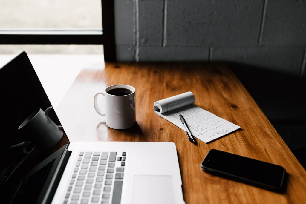 Laptop and mobile phone next to a notebook and coffe on a wooden desk