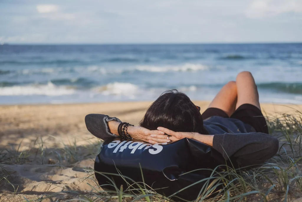 A girl resting on a beach with her hands behind her head and laying on a backapack