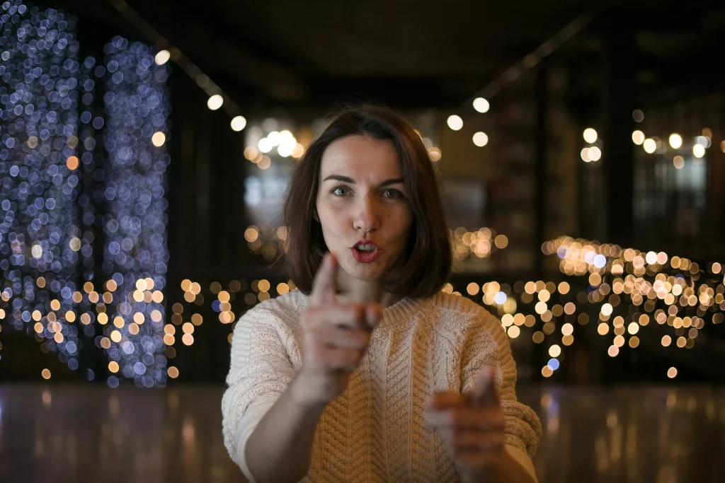 a women in a white shirt with lights in the background pointing at the camera