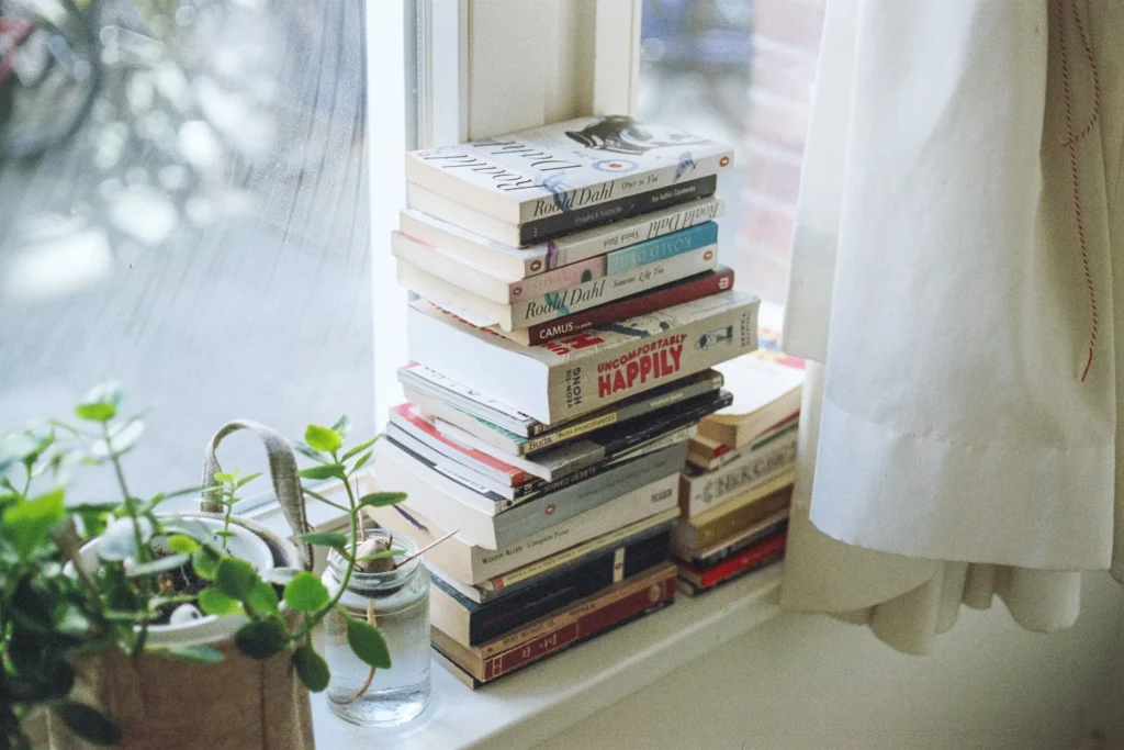 books stacked on top of each other next to a plant on a window behind a curtain