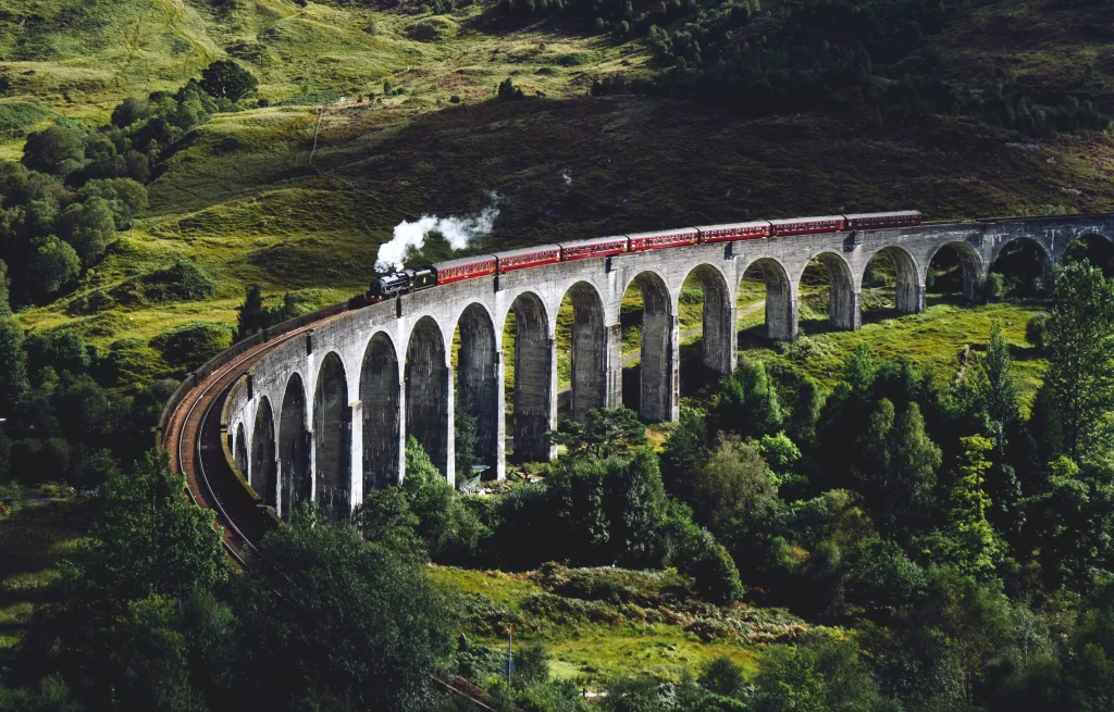 a red train passing through a green forest on a stone railway