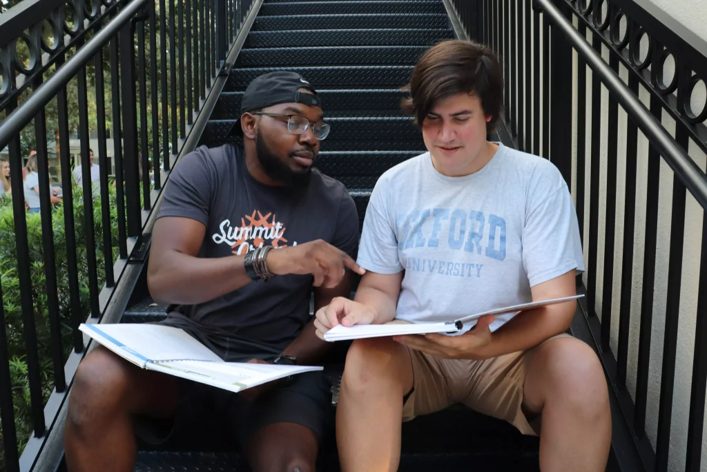 a guy in a white shirt and a guy with glasses siting on a stairway and reading a book together