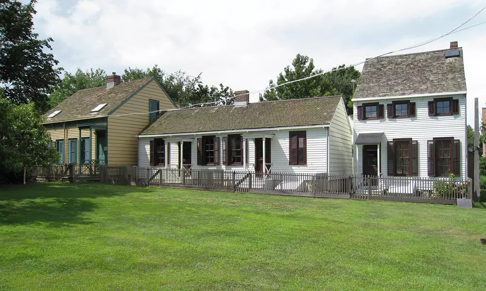 two white color houses and a yellow color house next to them. Infront is a green field